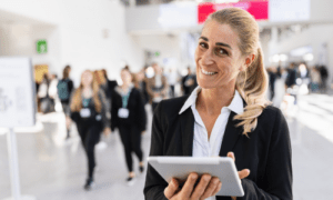 Freundliche Messehostess mit Tablet lächelt in die Kamera, im Hintergrund reges Messegeschehen mit vielen Personen – professionelles Personal auf Messen im Einsatz.
